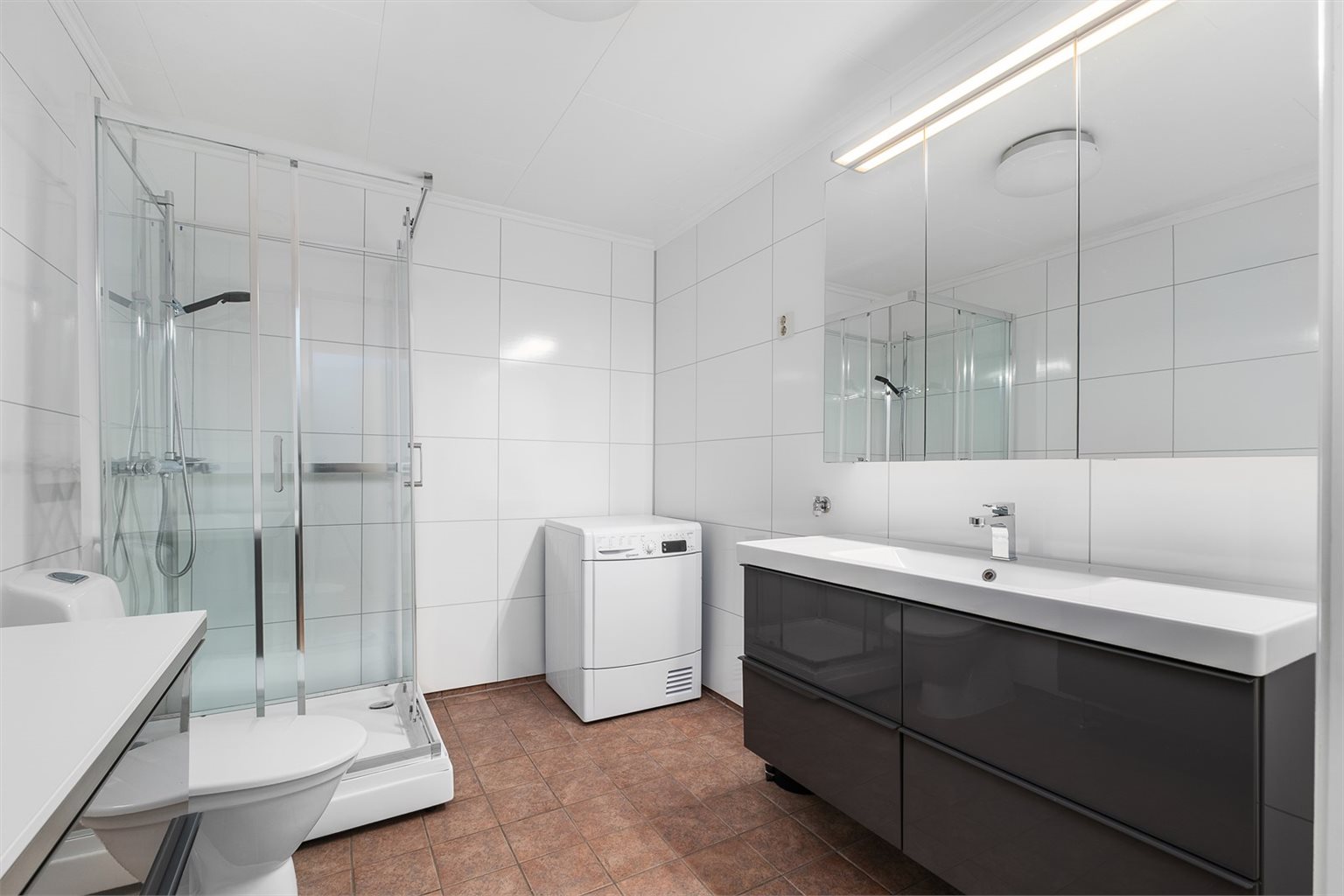 Badet er innredet med fliser på gulvet og baderomsplater på veggene, og har en funksjonell utforming. Her finner du dusjkabinett, toalett, servantskap med nedfelt servant og opplegg for vaskemaskin.