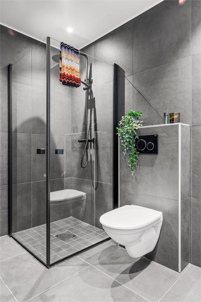Badet byr på et lekkert dusjhjørne med regnfallsdusj og innfellbare glassdører.  Baderomsinnredningen har heldekkende servant og skuffer under, samt et vegghengt speil med integrert belysning og rommet har vegghengt toalett.